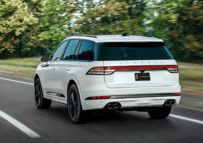 A white 2026 Lincoln Aviator® SUV is being driven away down a road lined with colorful trees. | Capital Lincoln of Wilmington in Wilmington NC