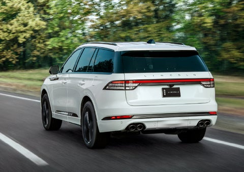 A white 2026 Lincoln Aviator® SUV is being driven away down a road lined with colorful trees. | Capital Lincoln of Wilmington in Wilmington NC