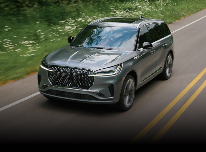 2027 Lincoln Aviator Reserve in Asher Gray, front three-quarter view driving on a winding tree-lined road.