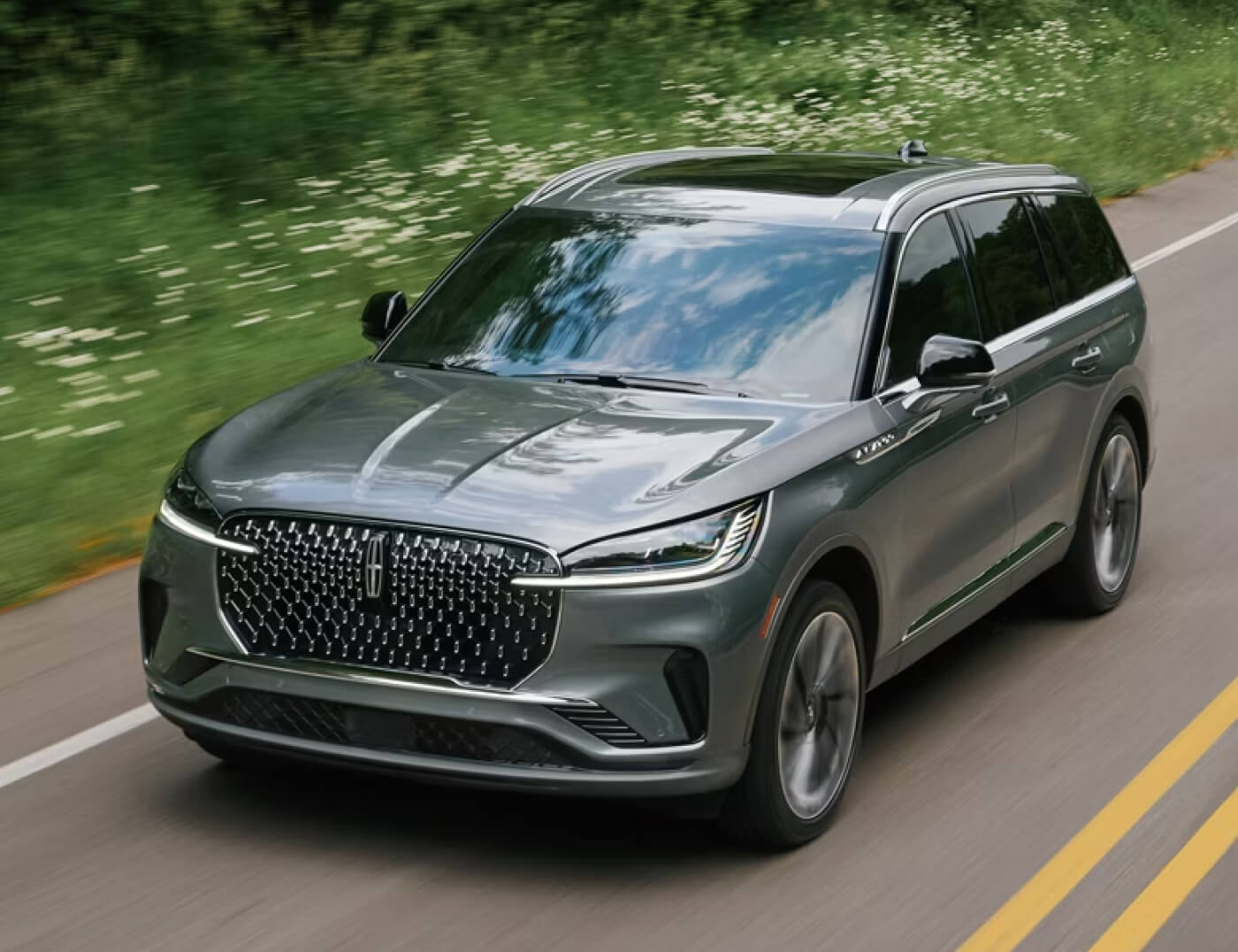 Front exterior action shot of a silver 2027 Lincoln Aviator driving on a forest highway.