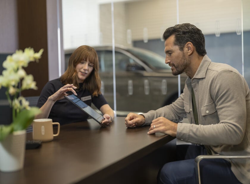 A Lincoln service advisor sitting at a desk and reviewing maintenance details on a tablet with a customer.